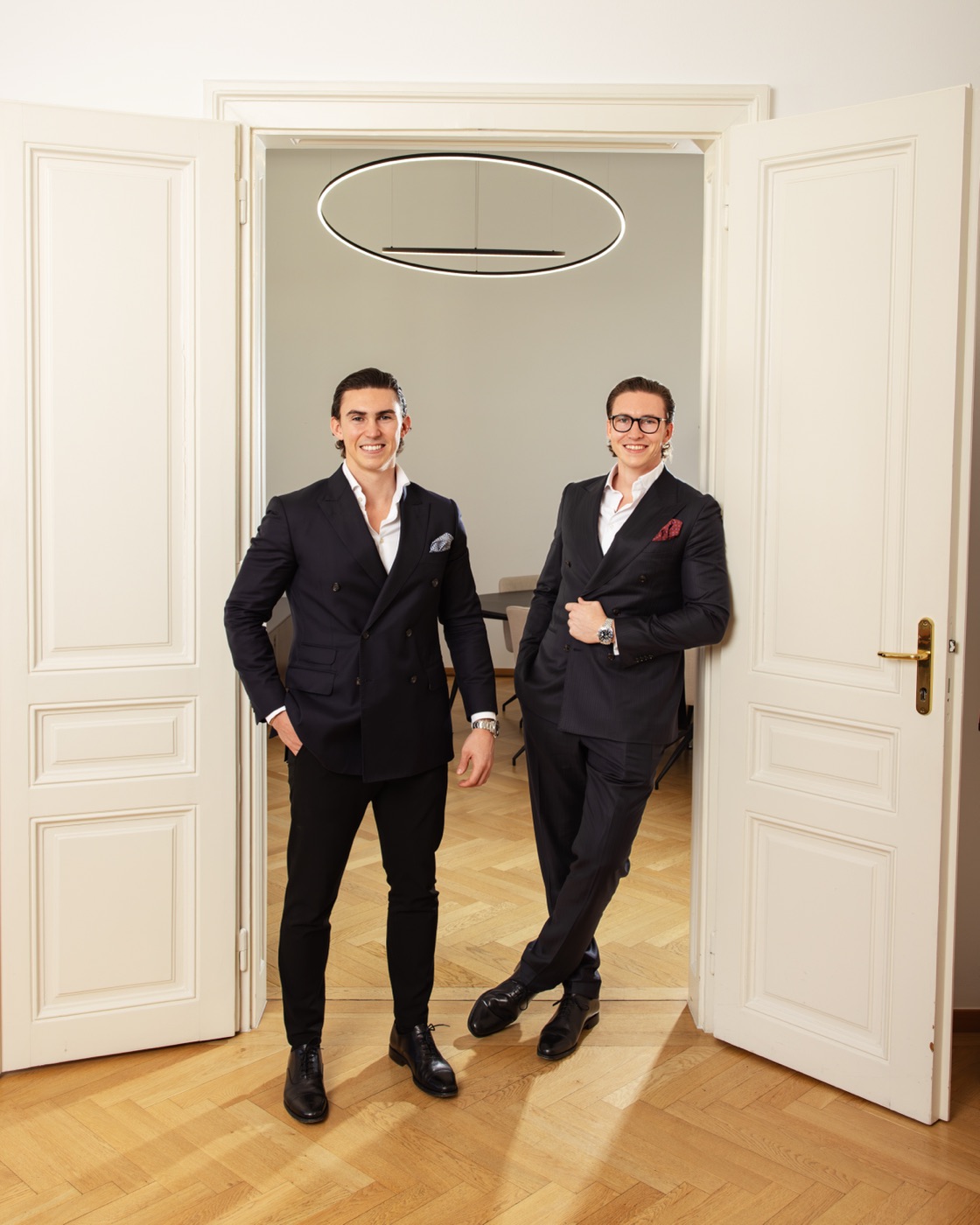 Jürgen and Matthias Nussbaumer in dark double-breasted suits smiling in a white period double door, a round pendant lamp and herringbone parquet behind them