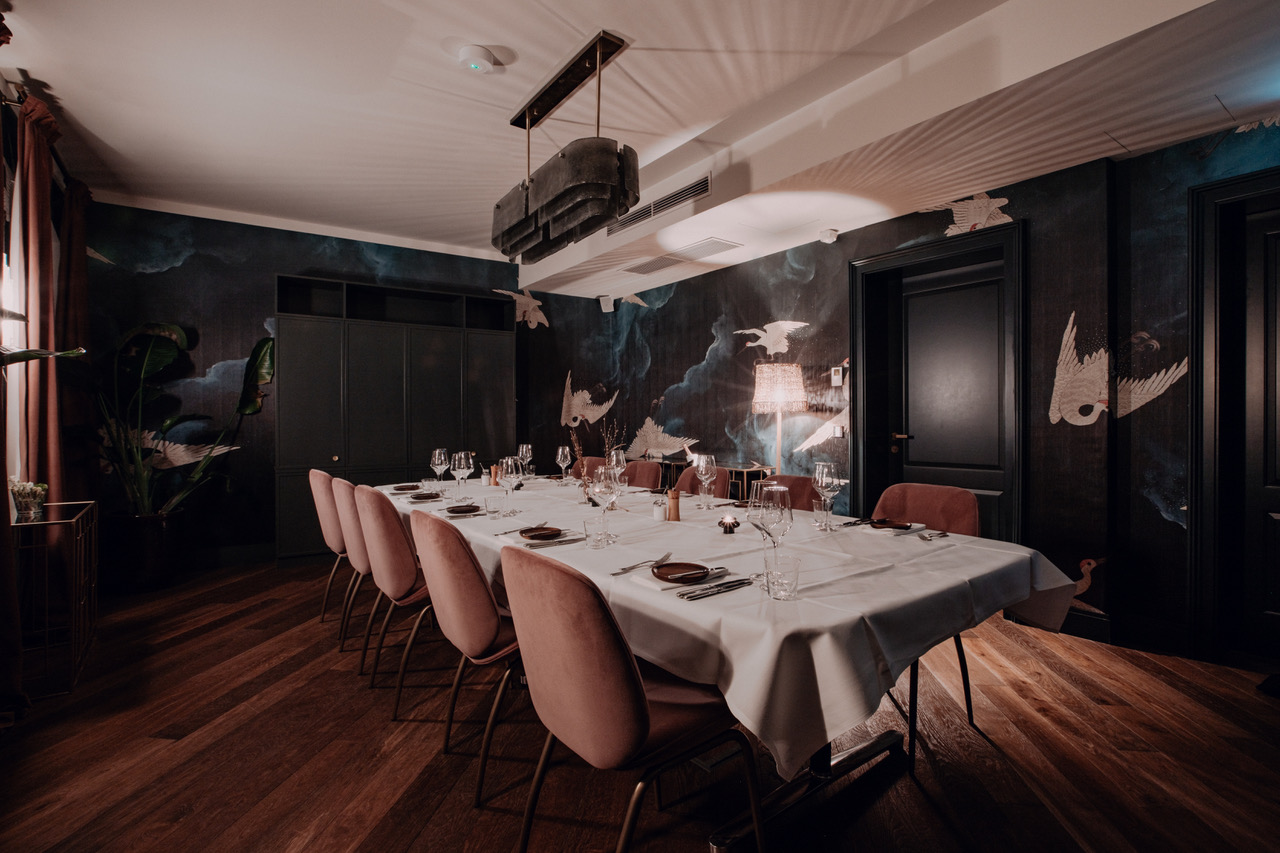 Long table laid with a white tablecloth and pink velvet chairs in a dark salon with painted birds on the walls