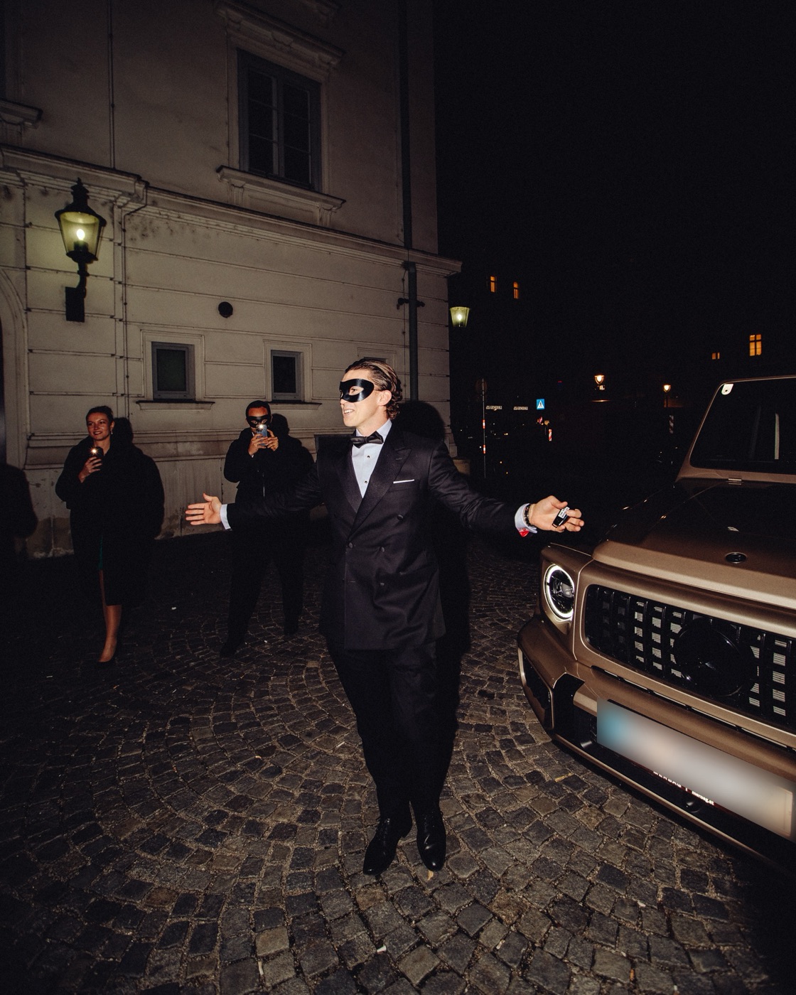 A man in a dinner jacket and black eye mask spreading his arms at night on a cobbled lane in front of a period building, an off-road vehicle beside him and guests with phones in the background