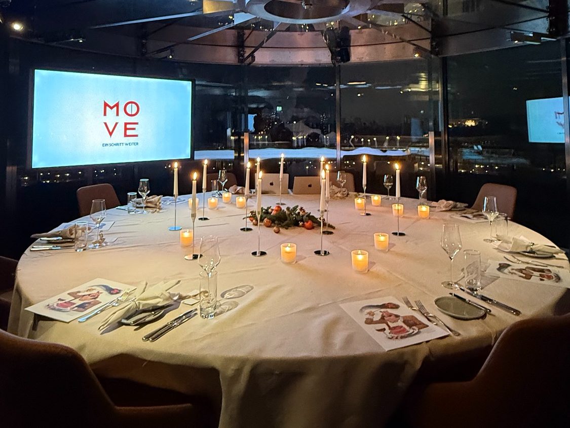 Round table festively laid with candles in a dark glass-walled hall above the city at night