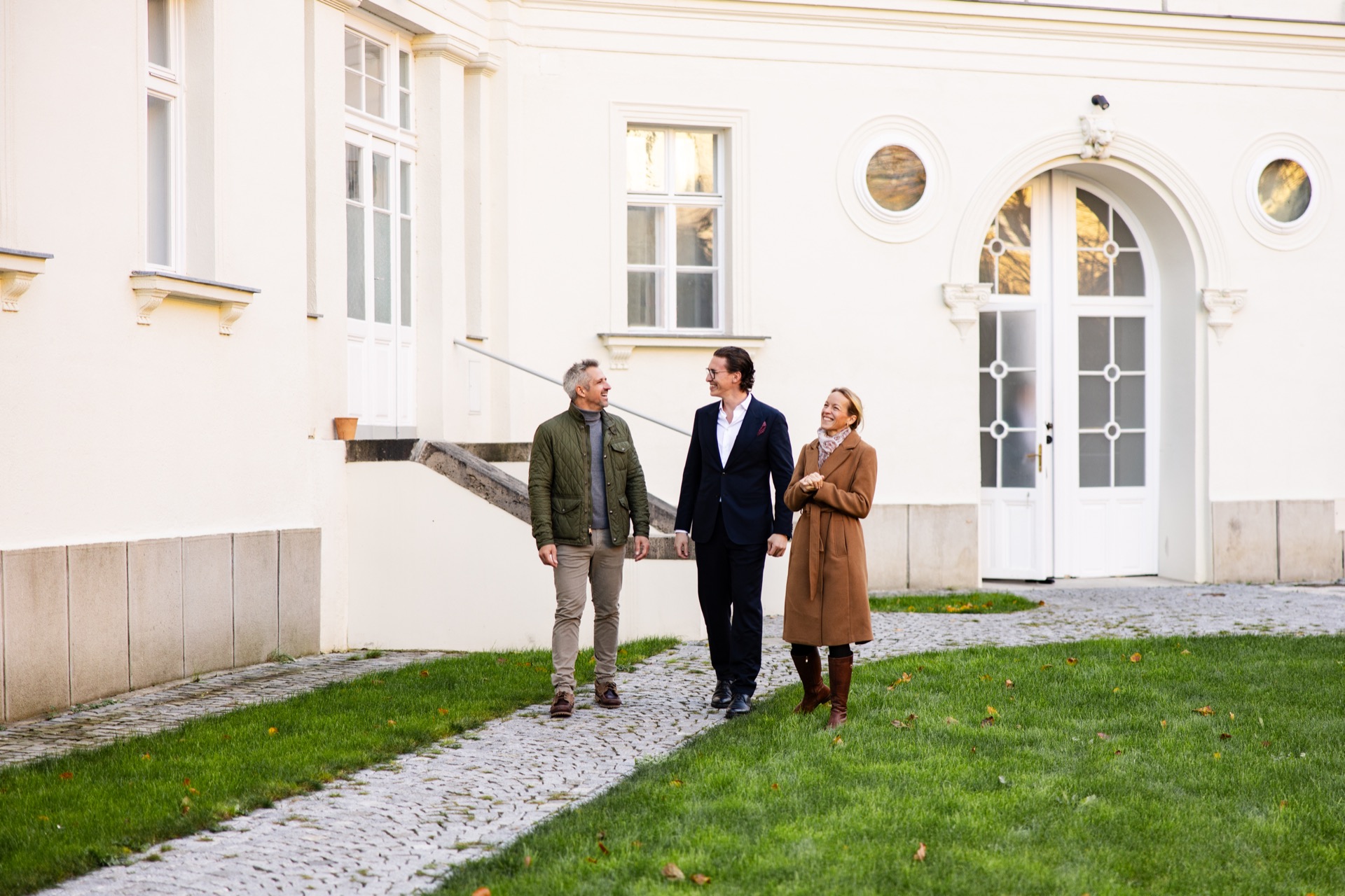 Jürgen Nussbaumer laughing as he walks with a man and a woman along a cobbled path in front of a white period building with an arched door