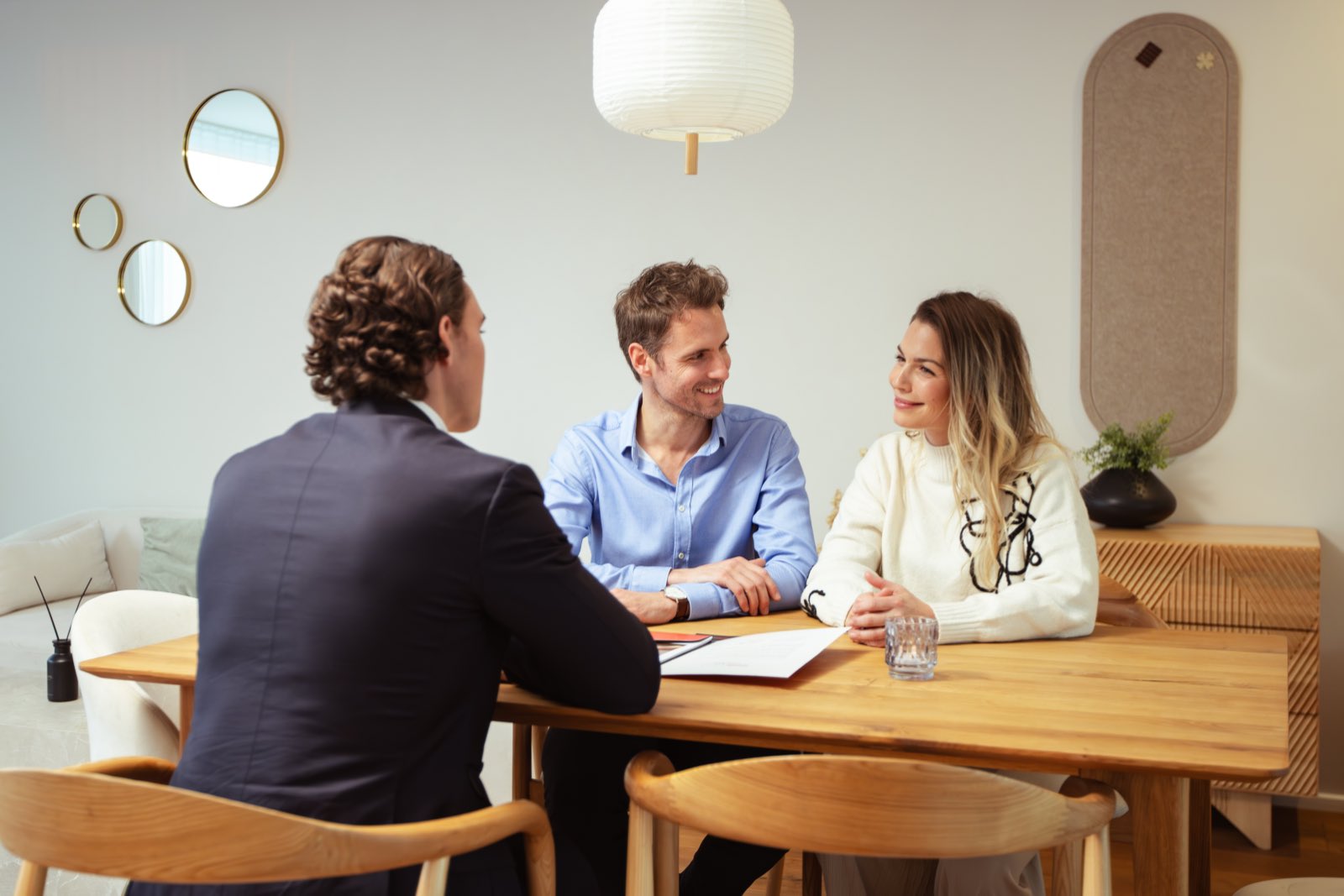 Advisory meeting at a wooden table: a consultant in a dark suit talking with a smiling couple in a bright living room