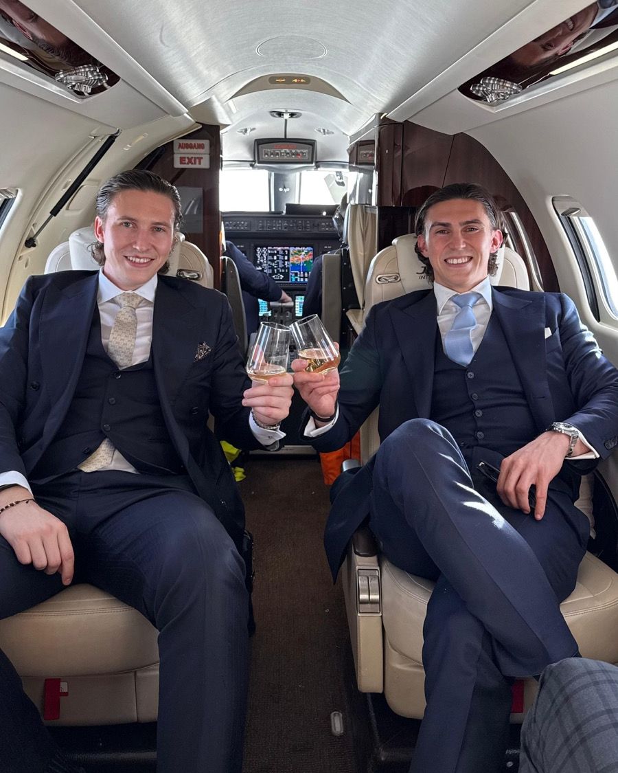 Jürgen and Matthias Nussbaumer in three-piece suits raising a glass in the cabin of a private jet on the way to Tuscany