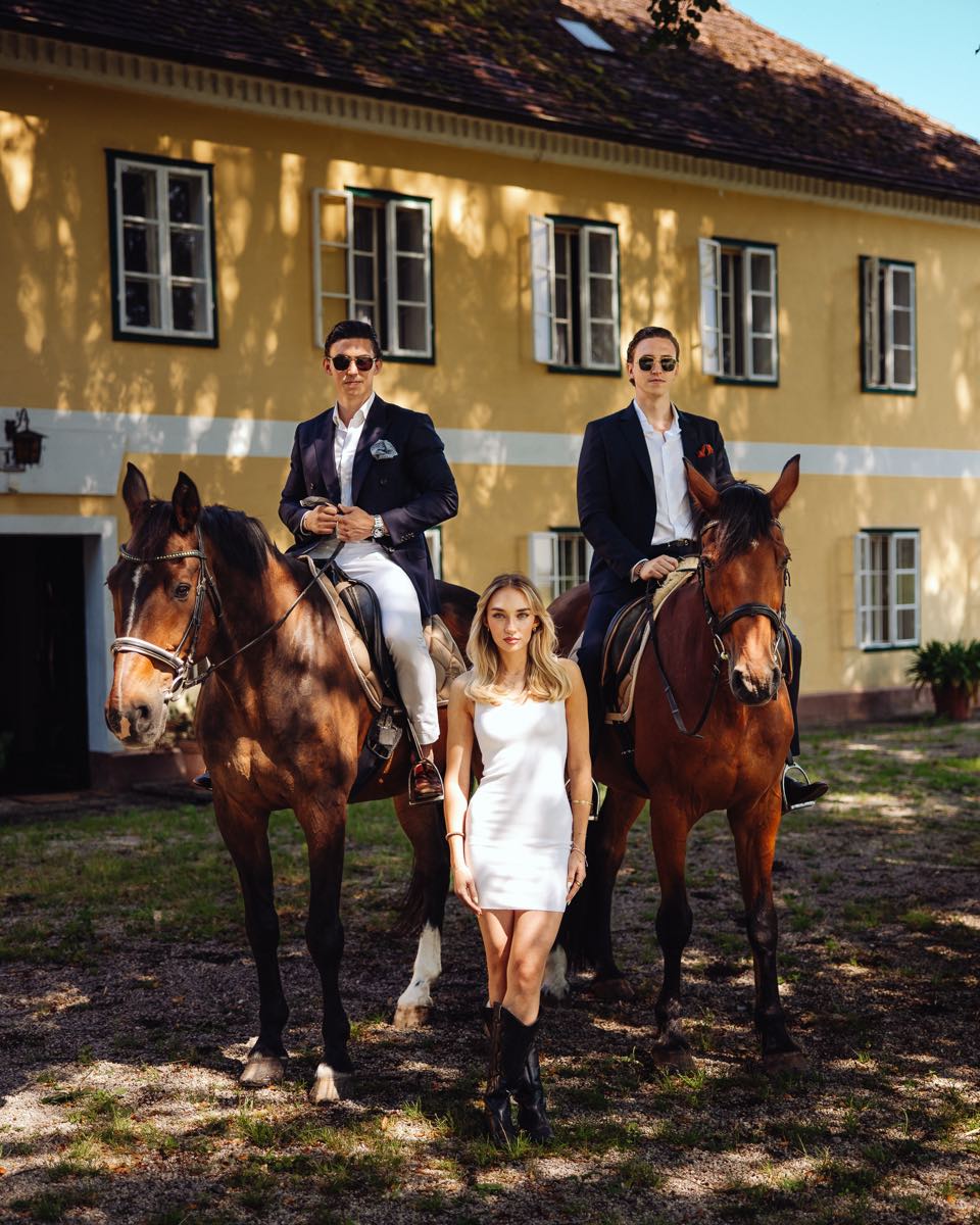 Two riders and a woman in a white dress in front of a yellow country house