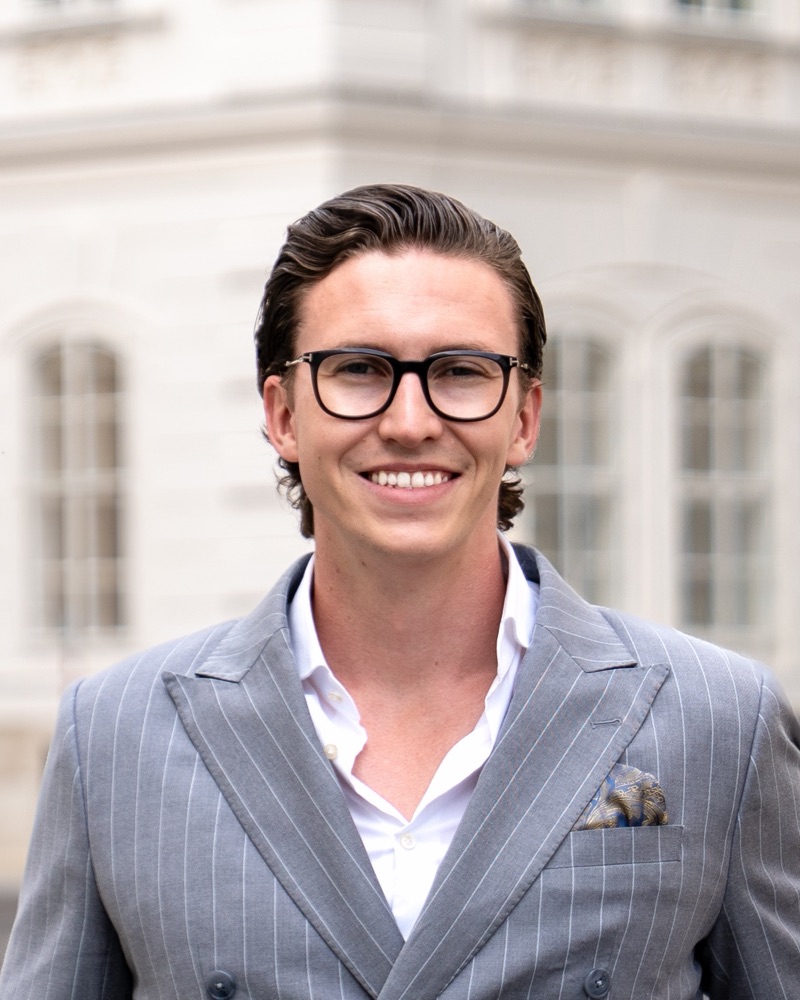 Portrait of Jürgen Nussbaumer with black glasses and a grey pinstripe double-breasted suit in front of a light period building in Vienna