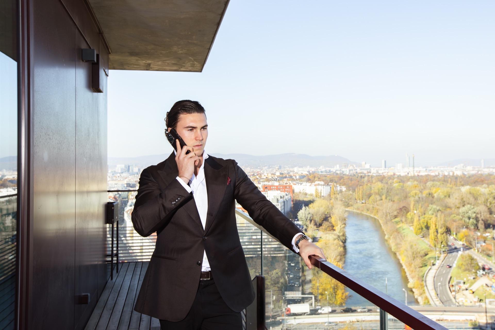 A man in a dark suit on the phone on a balcony high above the city, a river and autumn trees below