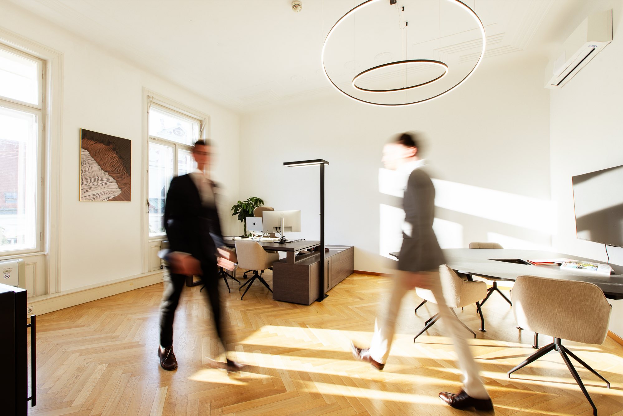 Two men in suits walking in motion blur through a bright period office with herringbone parquet and a round pendant lamp