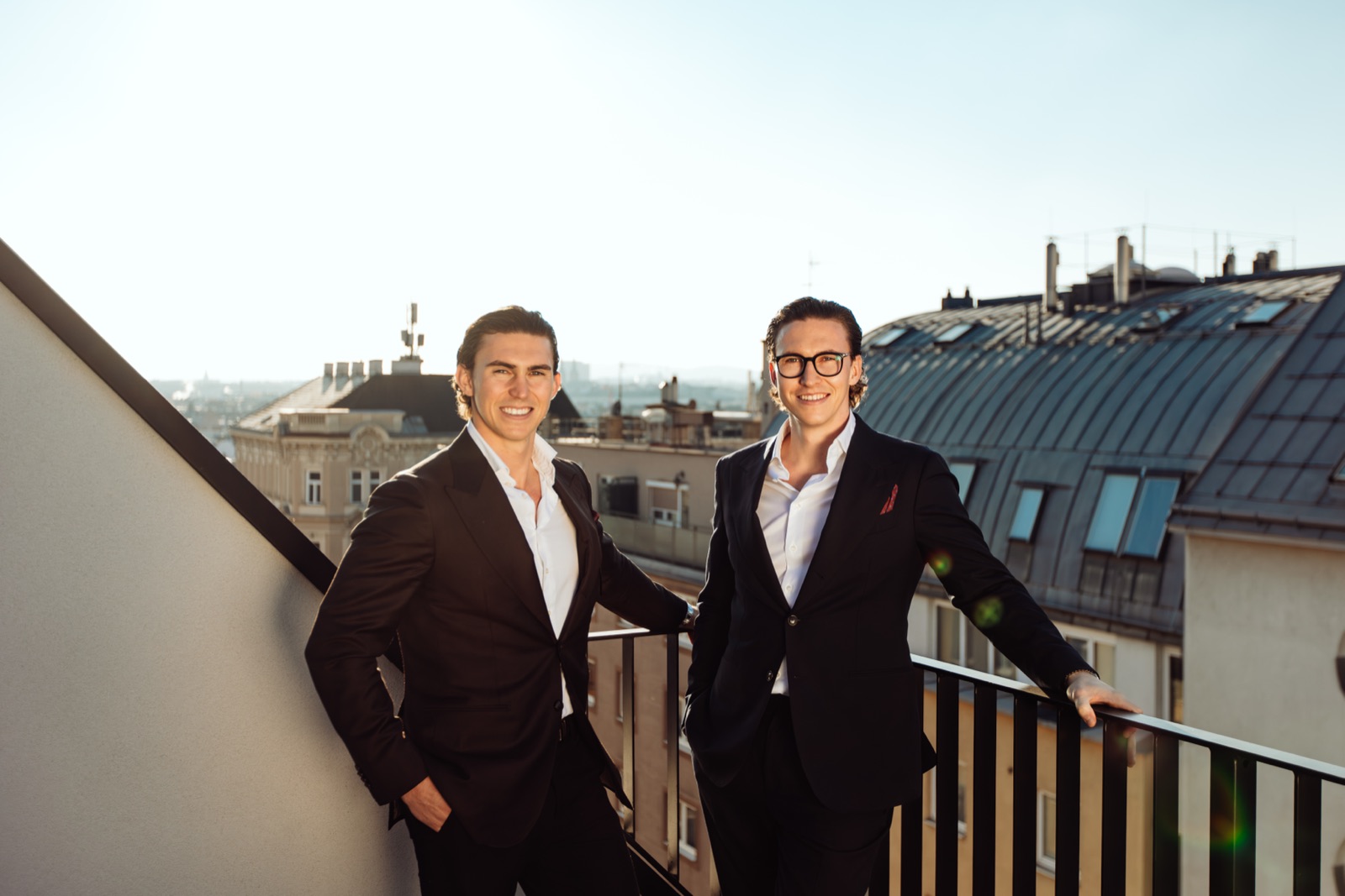 Jürgen and Matthias Nussbaumer in dark suits smiling on a rooftop terrace above the roofs of Vienna