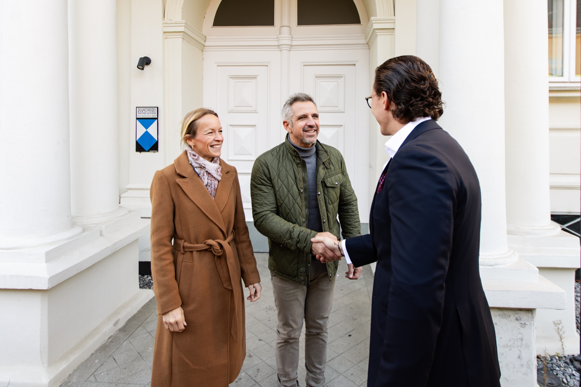 Jürgen Nussbaumer greeting a couple with a handshake at the entrance of a white period building with columns