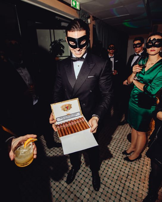Host in a dinner jacket and eye mask holding a cigar box
