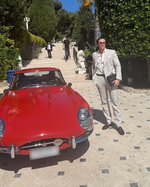 Man in a light suit next to a red classic car on an avenue lined with palm trees