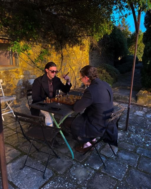 Two men playing chess in the evening sun on a stone terrace in front of an old country house